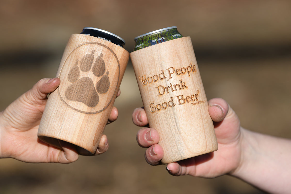 Two people clinking TreeSleeve wooden can coolers, featuring a paw print and “Good People Drink Good Beer” engraving.
